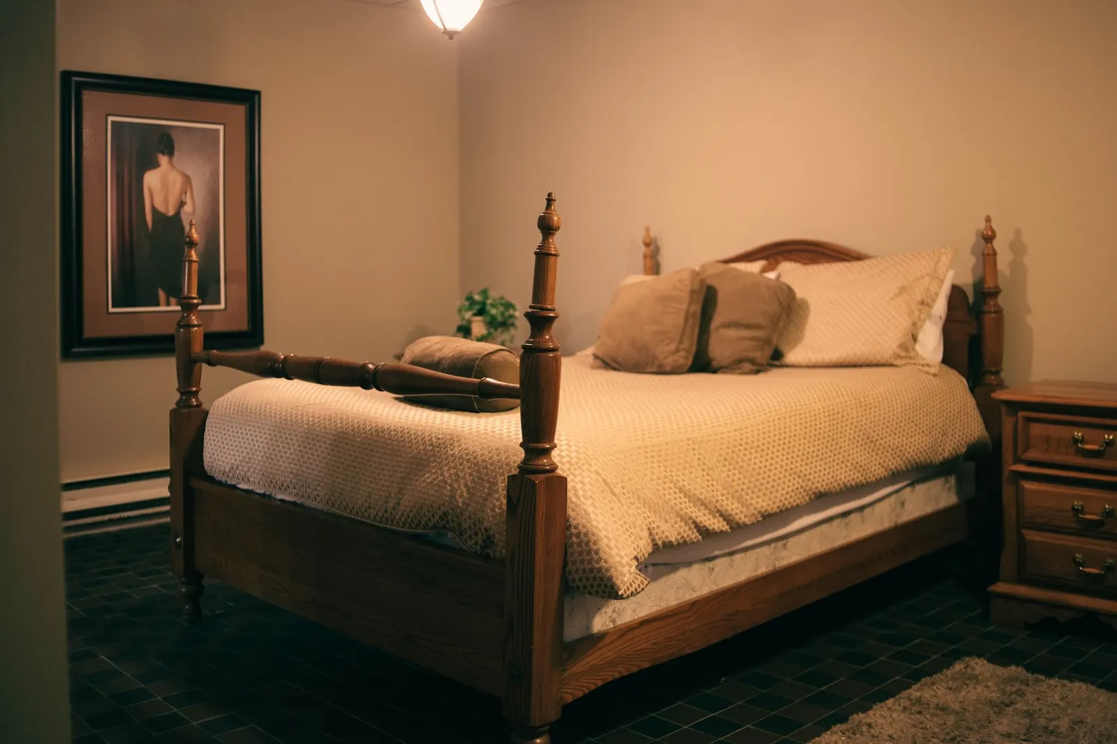 Warmly lit bedroom with wooden furniture, a made bed, and a classic painting on the wall.