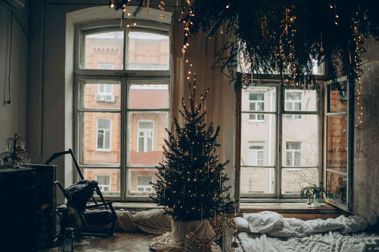 Small apartment living room with a slim lit Christmas tree in the corner and garland along a window