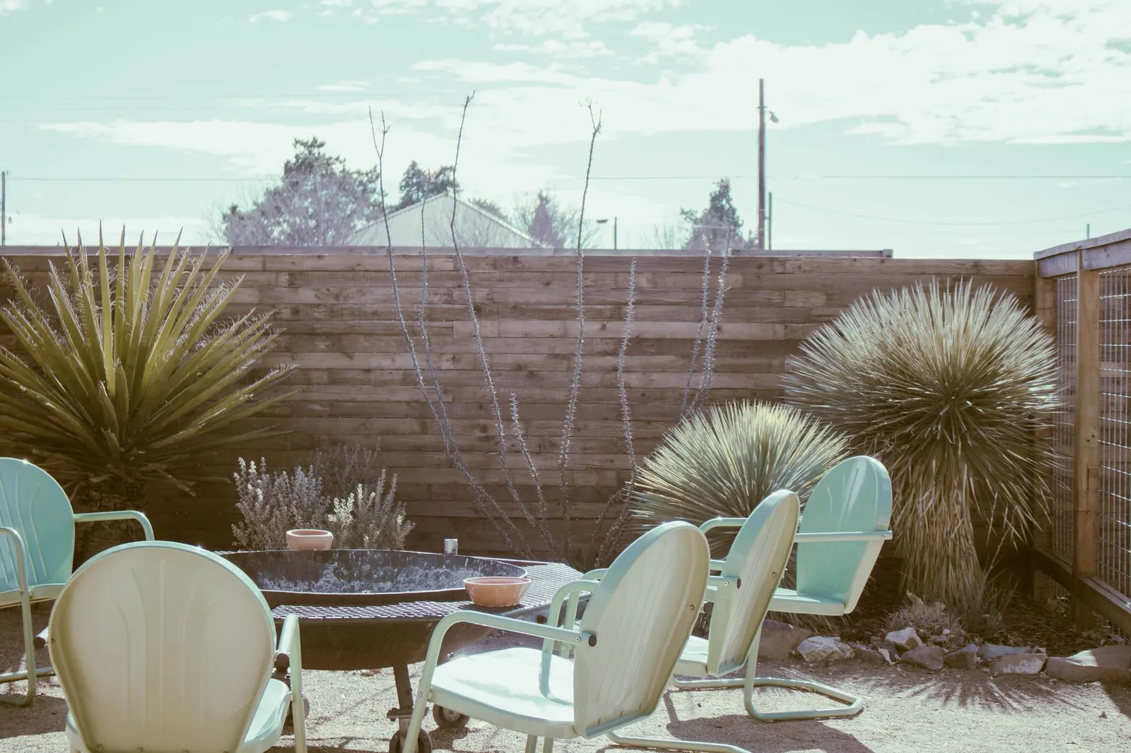 Desert patio with a shade sail, terra-cotta pots, a cactus and light cushions on metal chairs