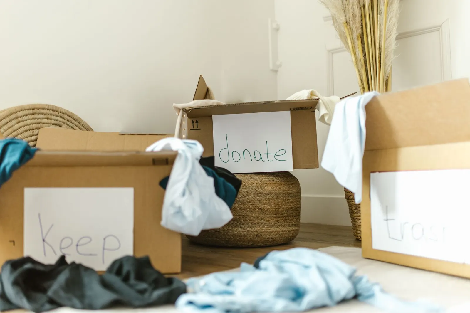 Clothes and household items sorted into labeled keep, donate and trash boxes on a wood floor