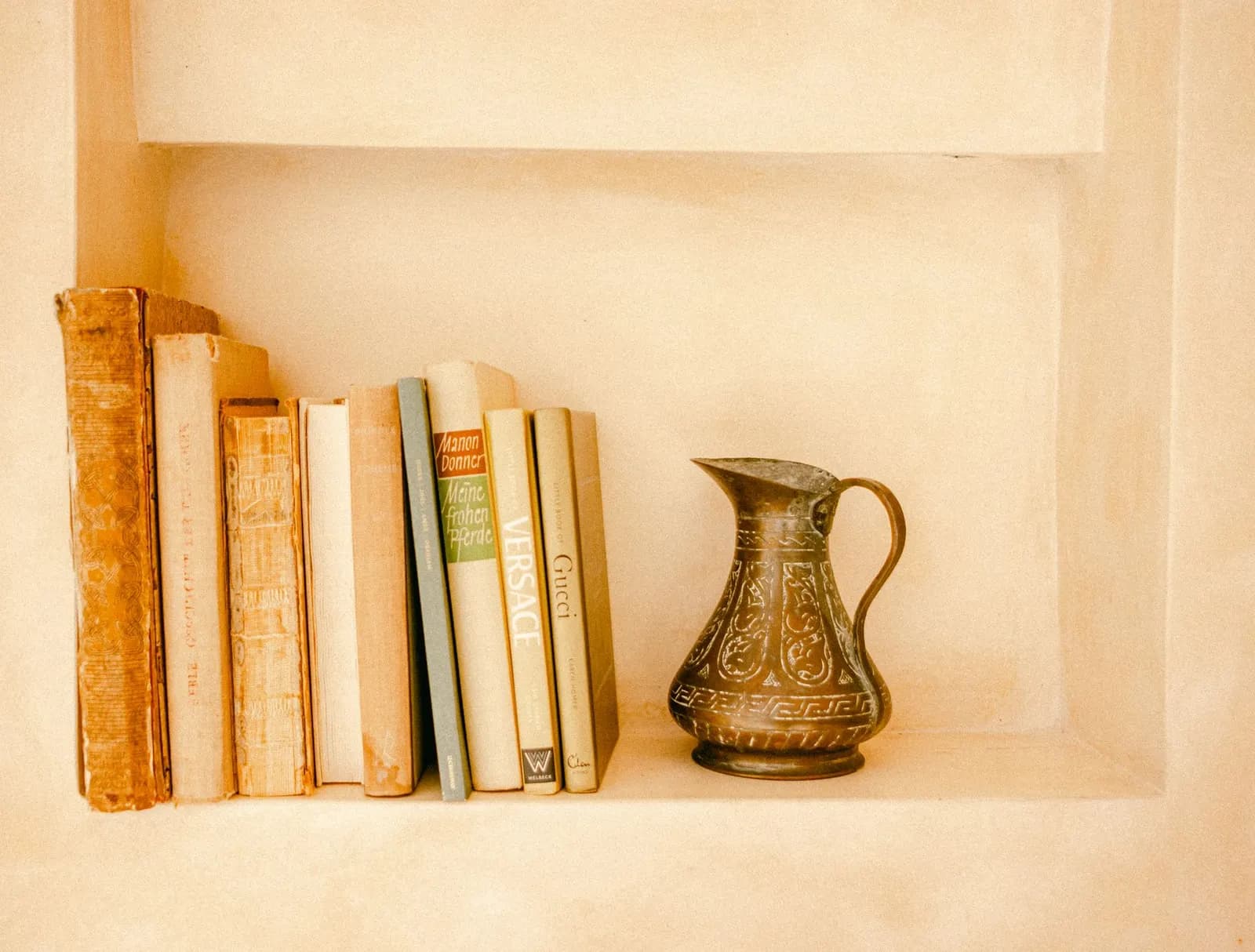 Aesthetic bookshelf with vintage books and an ornate brass jug in a warm-toned, minimalist setting.